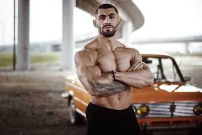 Portrait of shirtless muscular man standing by car on field