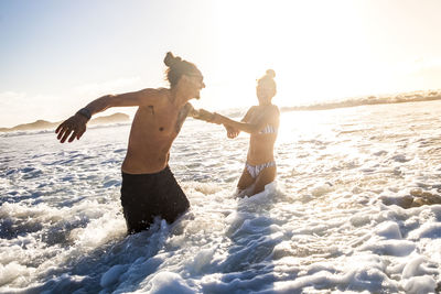 Girlfriend pulling boyfriend while standing in sea against sky during sunset