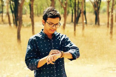 Young man looking at wristwatch standing against tree