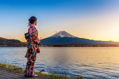 Woman standing by lake against sky during sunset