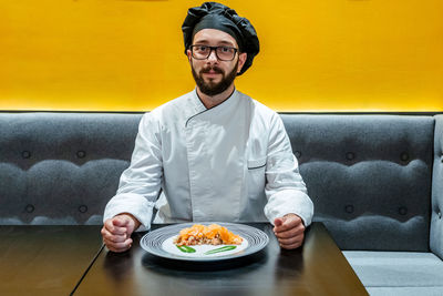 Content male cook in uniform sitting at table with tasty dish with fish and guacamole sauce while looking at camera