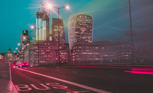 Light trails on city street by buildings against sky at night