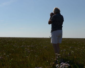 Rear view of senior woman standing on grassy field against sky during sunny day