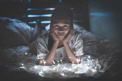 Close-up portrait of young woman lying by illuminated string lights on bed