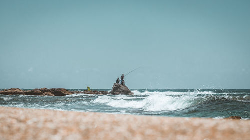 People fishing while sitting on rock at beach against clear blue sky during sunny day