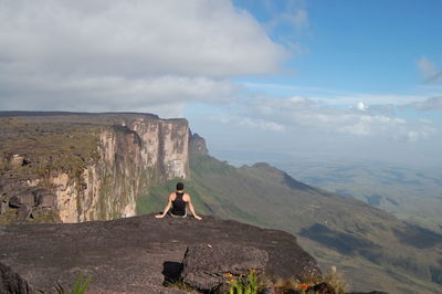 Man sitting on rock looking at mountain against sky