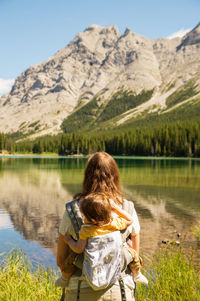 Mother carrying baby on carrier standing by a lake in the mountains on a sunny day