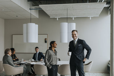 Smiling mature businesswoman standing by young businessman with coffee cup in board room