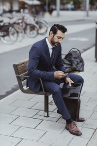 Entrepreneur with bag and file using smart phone while sitting on bench in city