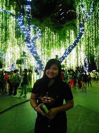 Portrait of smiling young woman standing by illuminated tree at night