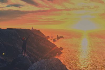 Rear view of woman with arms raised standing on rock at beach during sunset