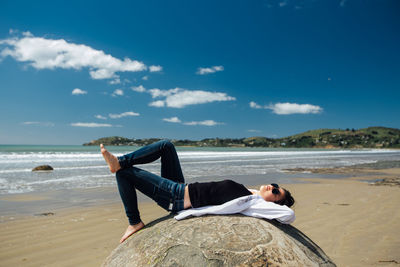 Side view of carefree woman lying on rock at beach against sky during summer