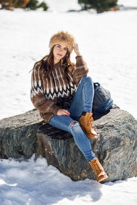 Full length portrait of young woman wearing warm clothing while sitting on rock amidst snow