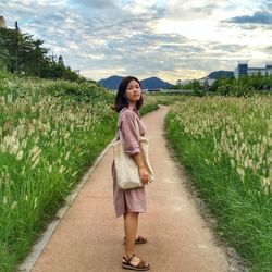 Young woman standing on footpath amidst grass growing against cloudy sky