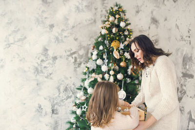 Mother and daughter decorating christmas tree against wall
