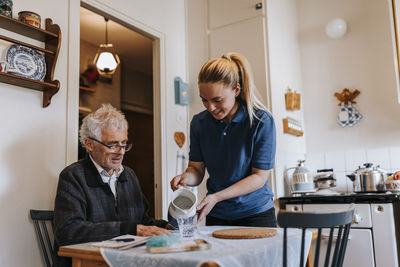 Female caregiver serving water to senior man sitting at dining table in home