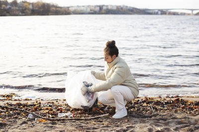 Young female environmentalist collecting waste while crouching by lake