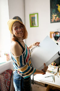 Side view of content female painter in funky outfit standing with art supplies in studio and looking at camera