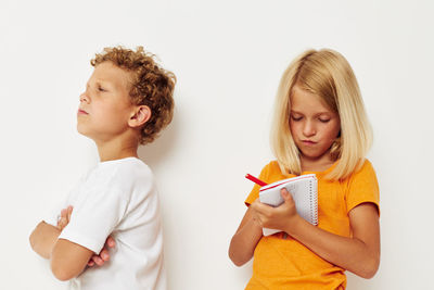 Girl writing on notebook while boy looking away against white background