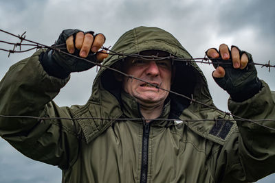 A young guy stands behind a barbed wire fence and holds onto the barbed wire with his hands.