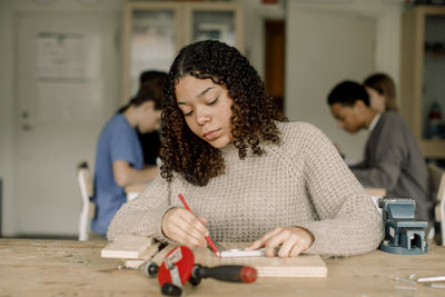 Concentrated female teenage student with curly hair marking on wood during carpentry class