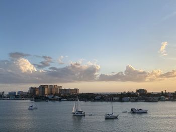 Sailboats in sea by buildings against sky during sunset