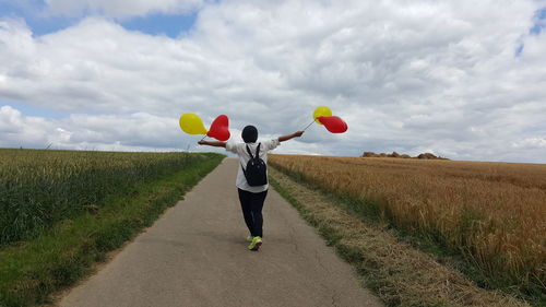 Rear view of woman with arms outstretched holding balloons while walking on road by field