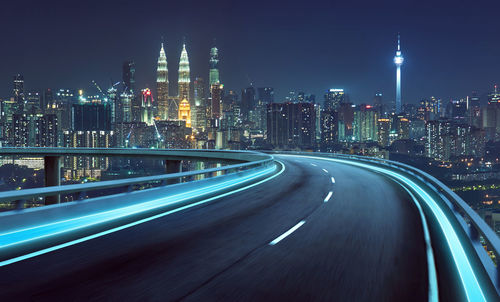 Light trails on road by illuminated buildings against sky at night