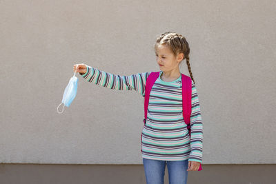A cute caucasian schoolgirl with a knapsack behind her back holds a medical mask