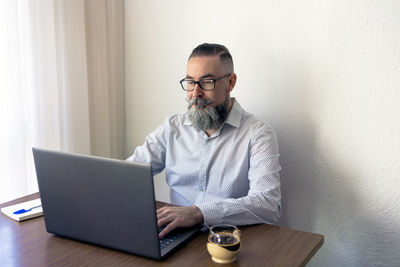 Bearded man with glasses having coffee and notepad at table working on laptop in home office
