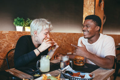 Cheerful woman and black male friend sitting in fast food cafe having fun while eating burgers