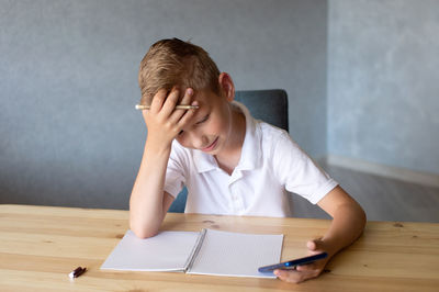 Cute boy writes in an open notebook and looks at the phone.