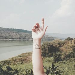 Cropped hand of person with tattoo by river against sky