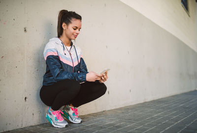 Full length of young woman using phone while crouching by wall