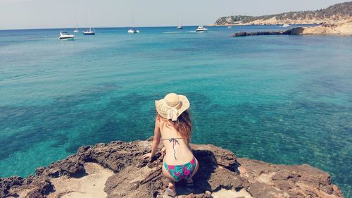 Rear view of woman wearing bikini while kneeling on rocks at beach