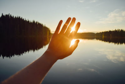 Cropped image of hand by lake against sky during sunset