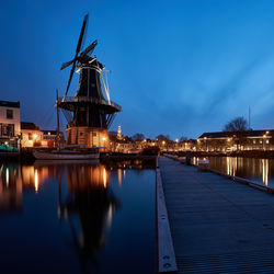 Old windmill and illuminated buildings by river against sky at dusk