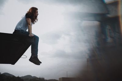 Low angle view of young man looking away while sitting on retaining wall against sky