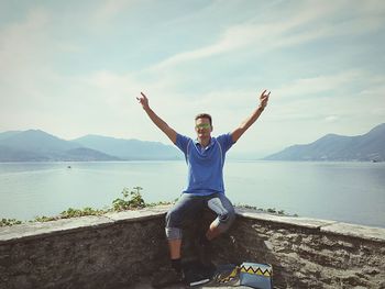 Portrait of man with arms raised sitting on retaining wall with lake in background 