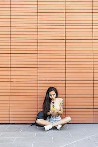 Young woman using mobile phone while sitting against wall