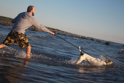 Side view of young man with dog running in sea against clear sky during sunset