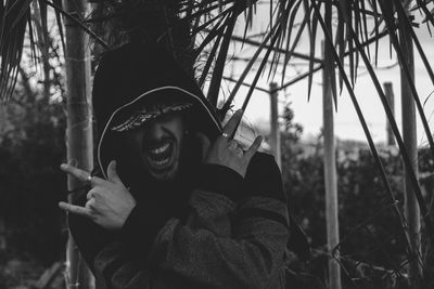 Close-up of man in hood clothing gesturing horn sign against trees