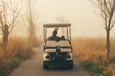 Rear view of father with son in golf cart on road amidst field during foggy weather