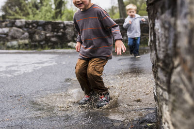 Midsection of boy jumping in puddle with brother in background