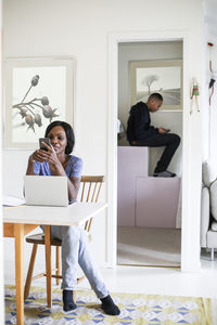 Mid adult woman using mobile at table with son sitting in storage room