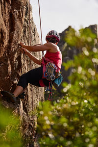 Low angle full length of active senior woman climbing on rocky cliff against sky