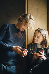 Smiling male and female friend using mobile phone while sitting at home