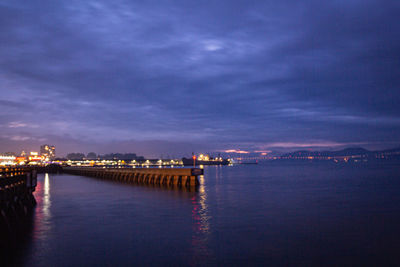 Illuminated city by sea against sky at dusk