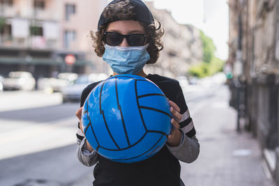 Portrait of boy wearing sunglasses holding ball while standing on street