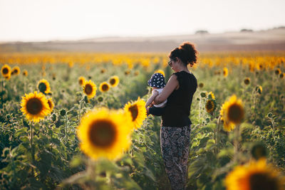 Side view of mother carrying daughter while standing amidst sunflowers in field against sky